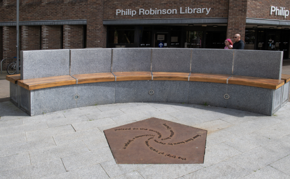 A bronze pentagram embedded into the floor before a curved bench. Five poetic lines swirl into the centre of the pentagram